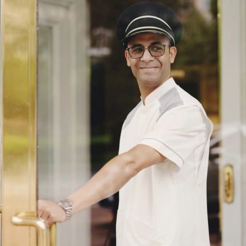 A person wearing a cap and uniform is smiling and holding a door open.