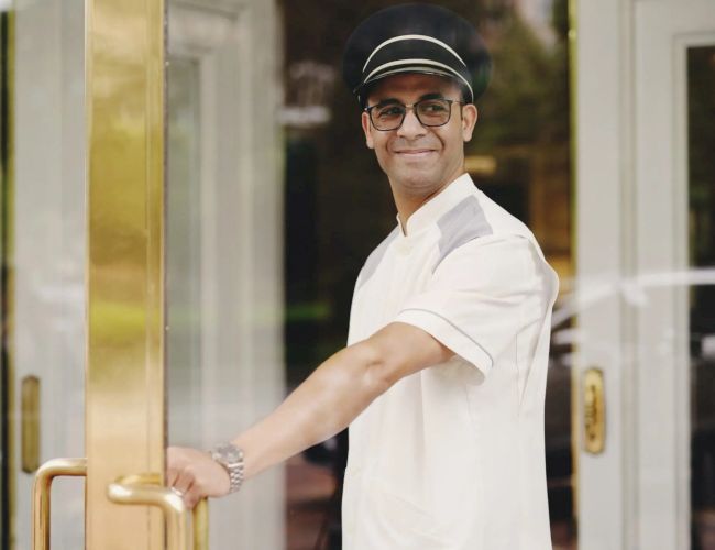 A person wearing a cap and uniform is smiling and holding a door open.