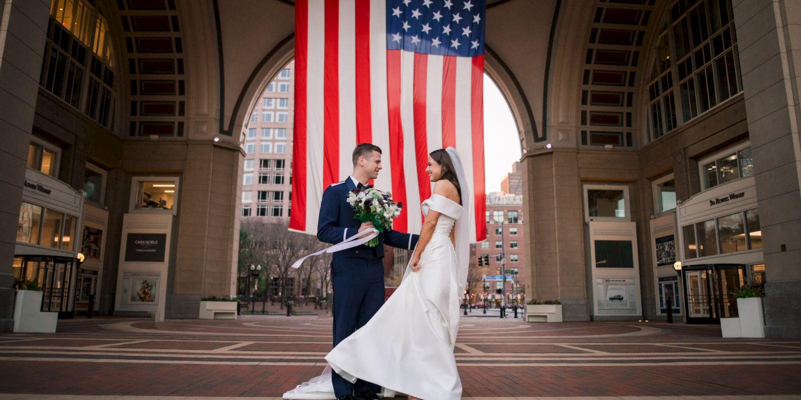 A couple in wedding attire poses under a large American flag in an open building atr