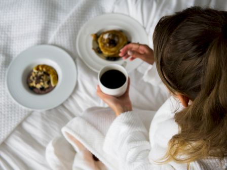 A person in a white robe enjoys breakfast in bed with coffee and pastries.