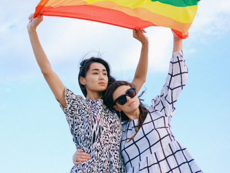 Two people are holding a rainbow flag against a blue sky.