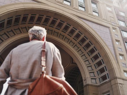 Person with grey hair and a shoulder bag walking towards an arched building entrance.