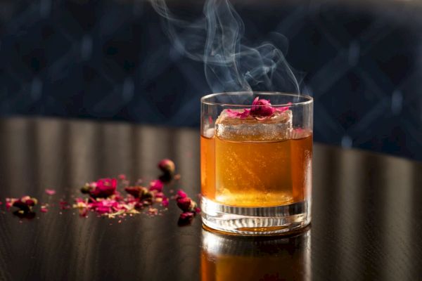 A smoky cocktail with ice and a flower garnish, surrounded by petals.