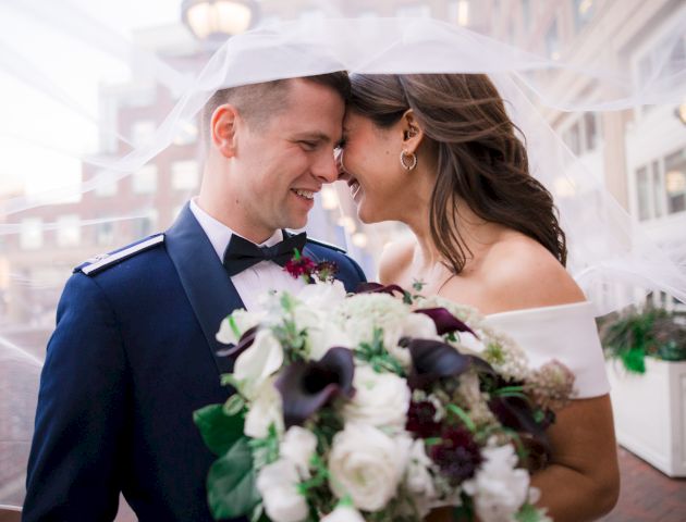 A bride and groom smiling at each other, with a bouquet and veil.