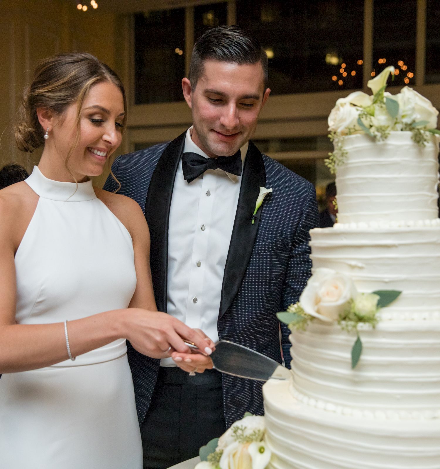 A couple is cutting a wedding cake together, smiling.