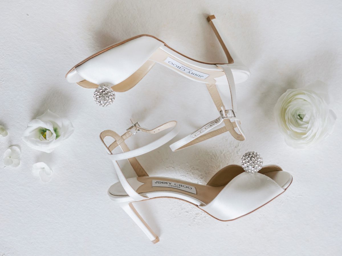 A pair of elegant white high-heeled shoes with embellishments lies beside flowers.