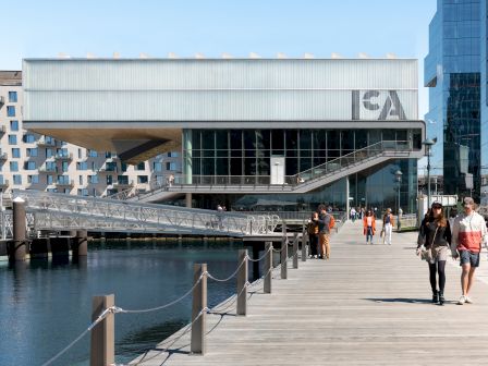 Modern building by a waterfront with people walking on a sunny day.