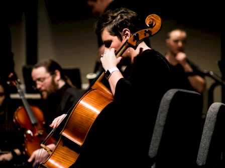 A musician plays the cello on stage, surrounded by fellow orchestra members.