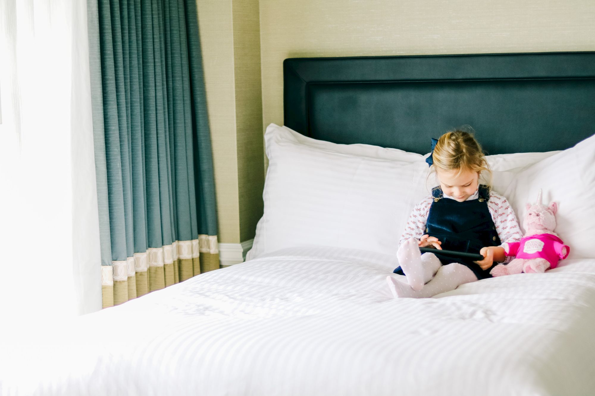 A child is focused on a tablet on a white bed.