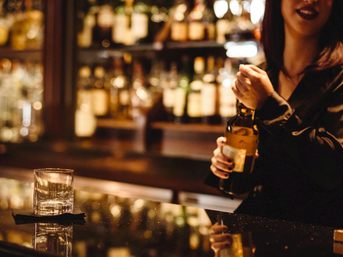 A bartender behind a bar counter holds a bottle, with a glass and black coaster on the counter, and shelves of liquor bottles in the background.