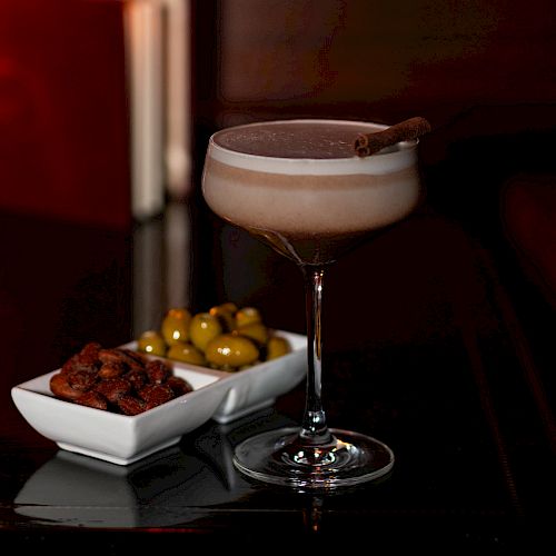A cocktail in a stemmed glass beside olives and snacks, set on a dark background.