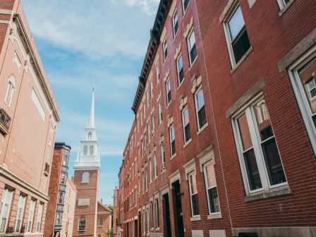 Urban street lined with brick buildings and a church spire in the distance against a