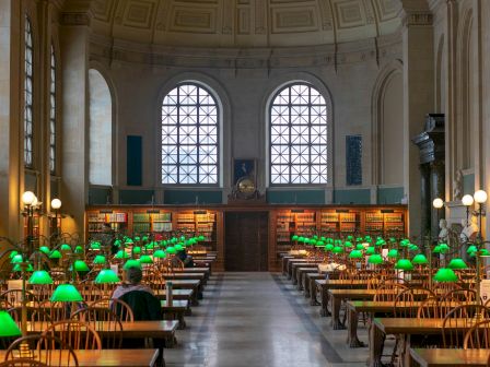 An elegant library reading room with rows of desks, green lamps, and large windows