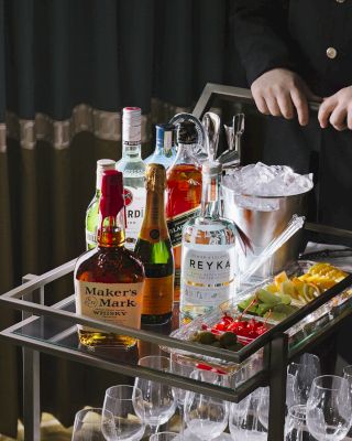 A person stands behind a cart with assorted liquor bottles, glasses, ice, and