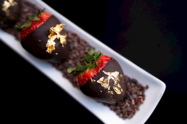 Three chocolate-covered desserts with strawberries and gold leaf on a plate, presented elegantly