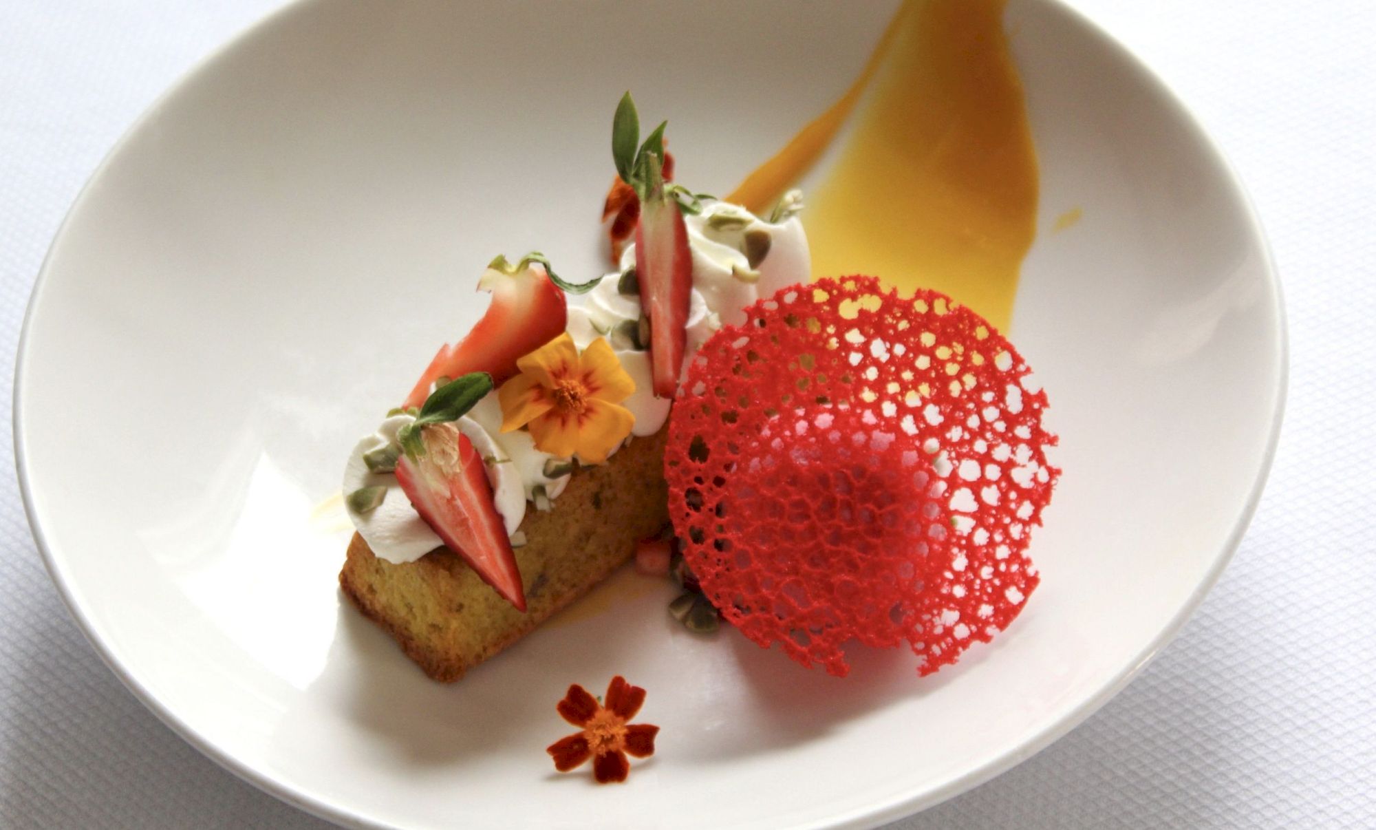 A dessert with cake, cream, fruit, edible flowers and a crisp tuile.