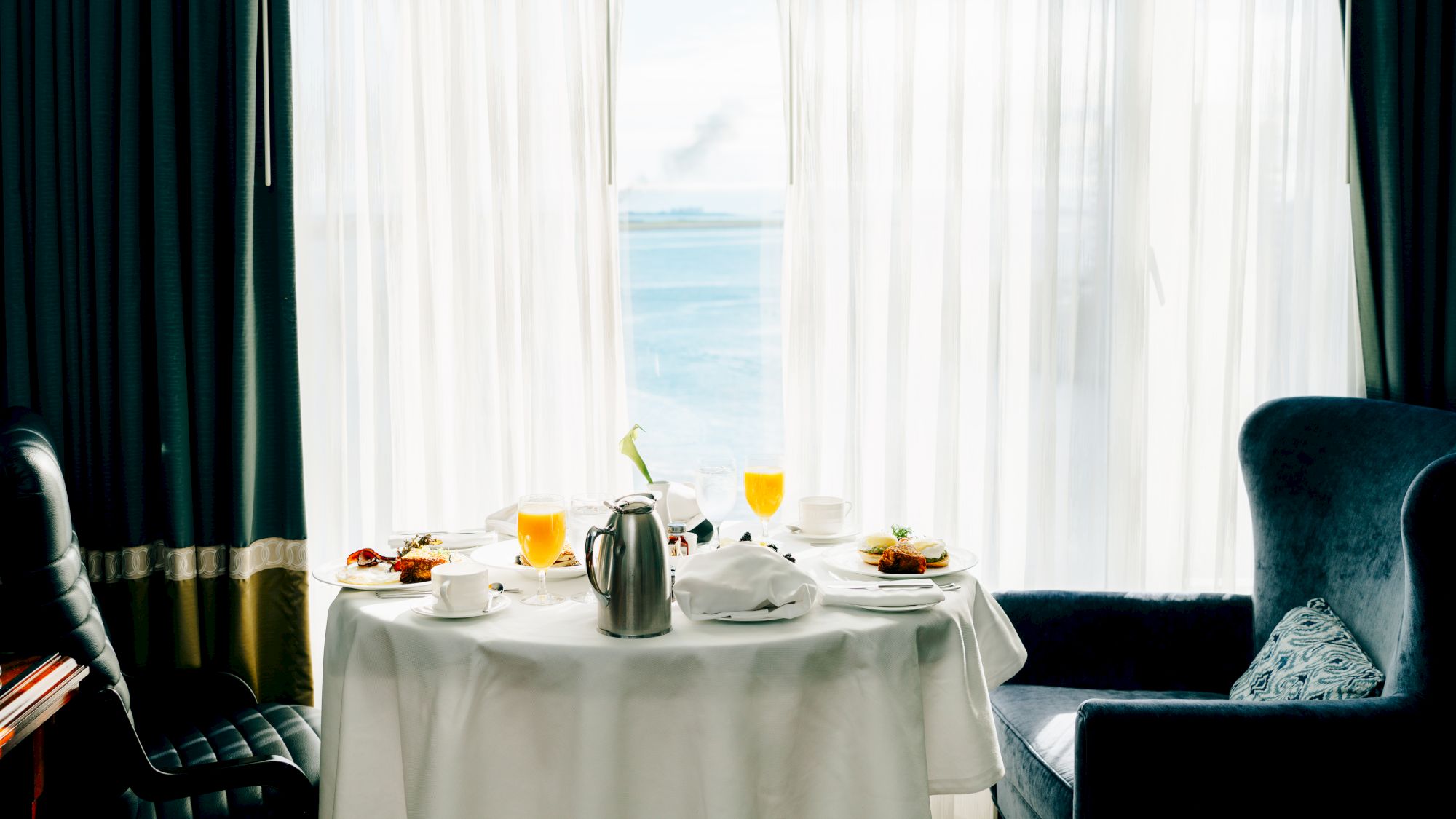 A table set for two with breakfast, a view of the ocean through curtains.