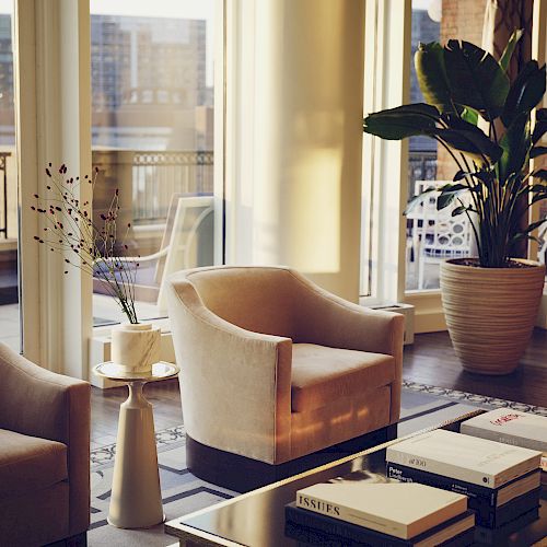 Cozy room with chairs, table, books, and a plant near sunny windows.