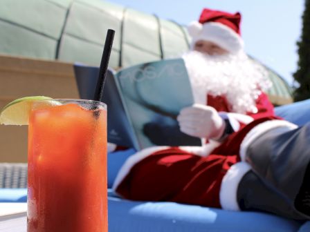 Santa Claus relaxing on a lounge chair, reading a magazine with a cocktail garnished with a lime wedge in the foreground.