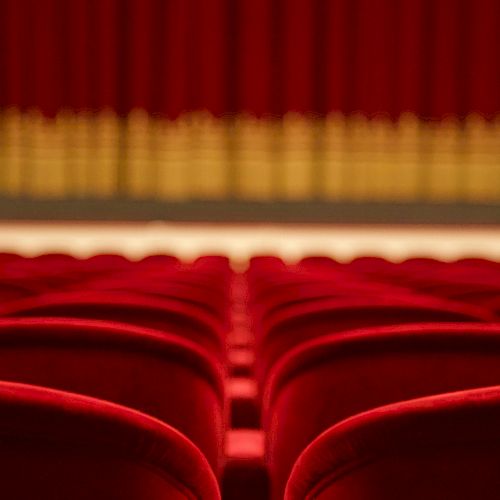 The image shows rows of red theater seats facing a stage with a closed curtain in the background, creating a classic theater ambiance.