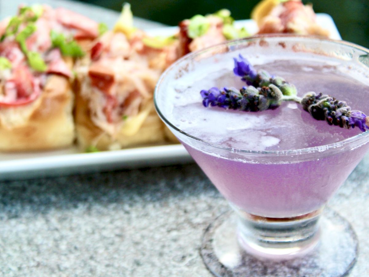 A lavender cocktail garnished with herbs in the foreground; lobster rolls in the back.