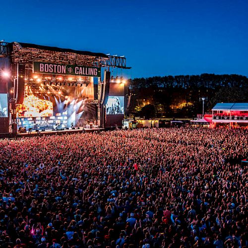 A large outdoor concert with a huge crowd and a stage lit up with colorful lights at night. The event is named 