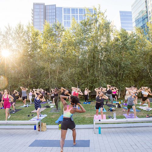 A group of people is participating in an outdoor fitness class on yoga mats in a park with a cityscape in the background, at sunset.