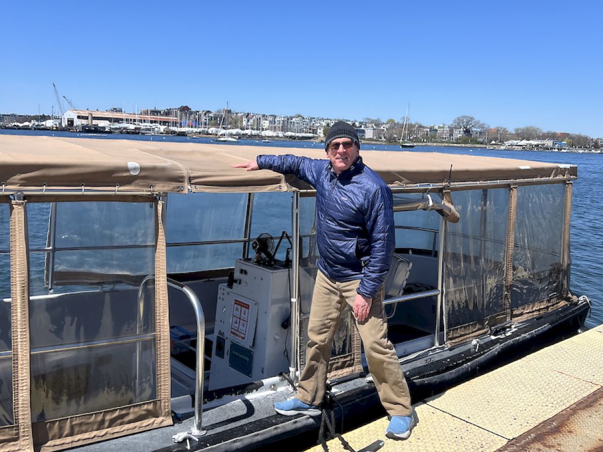 A person stands on a dock beside a boat with a tarp rooftop, next to a body of water; clear sky in the background.