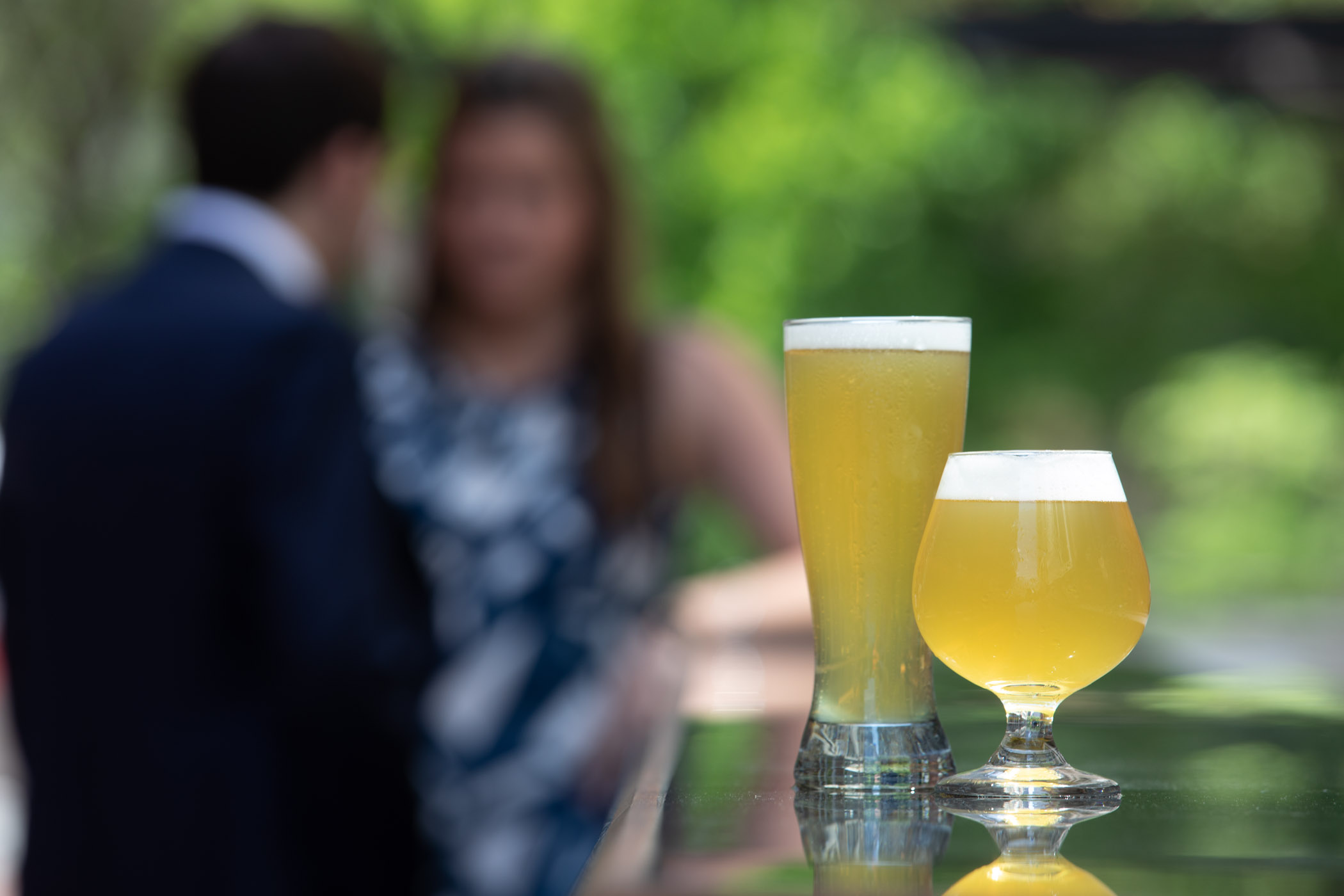 Two glasses of beer in focus with blurry people in the background.