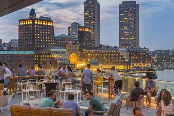 An evening at a rooftop bar with people socializing, overlooking a cityscape and waterfront.