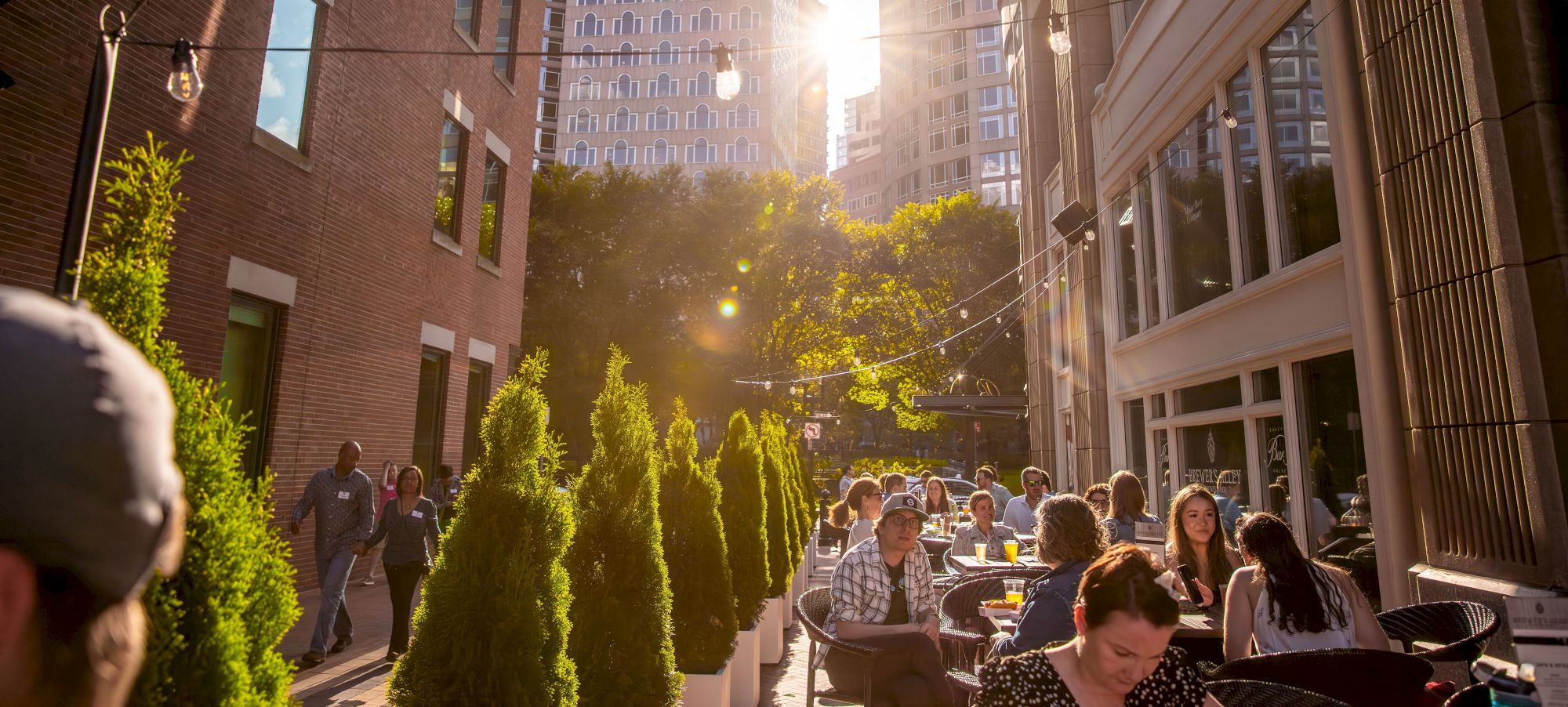 People dining outdoors, sun shining through buildings, urban setting.