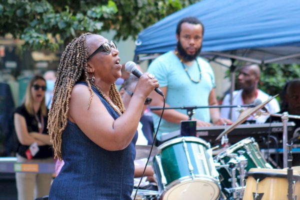 A woman is singing into a microphone with a band performing behind her. Others watch the performance outdoors under a canopy.