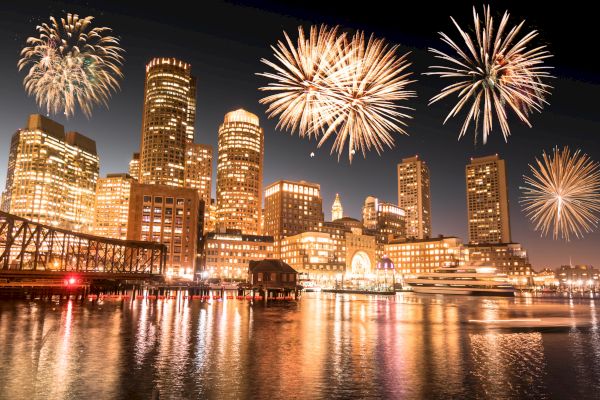 A cityscape at night with fireworks lighting up the sky, reflecting off the water. High-rise buildings and a bridge are prominently visible.