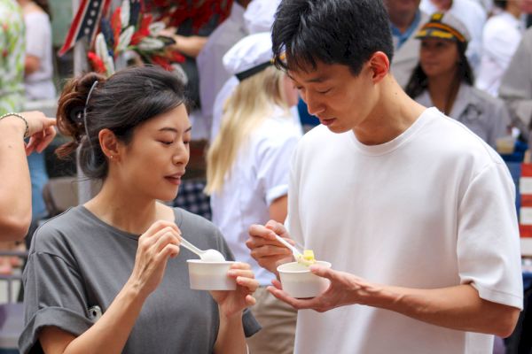 Two people enjoying food from small bowls, surrounded by others in what appears to be an outdoor event or market.