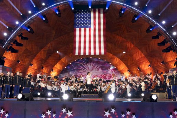 A large American flag hangs above a stage where an orchestra is performing. The venue is decorated with lights and star-themed decor, ending the sentence.