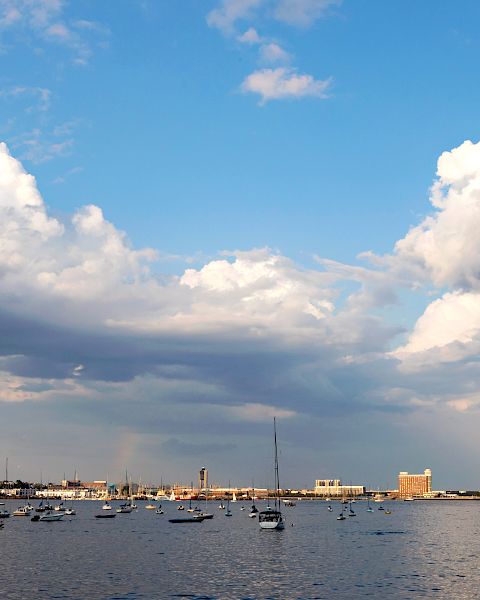 A serene waterside view featuring numerous boats, a distant shoreline with buildings, and a sky filled with large, fluffy clouds ending the sentence.