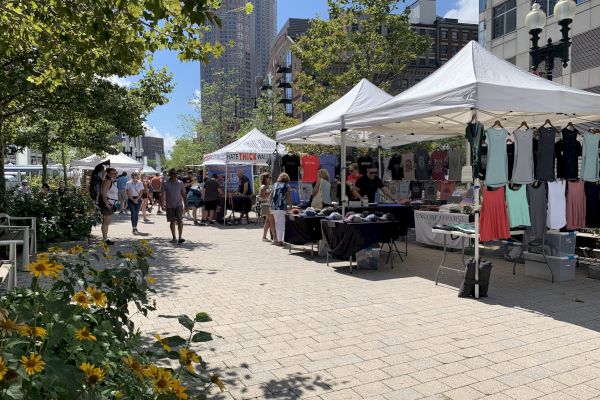 The image shows an outdoor market with several white tents, each selling different items such as clothes. People are walking around, and flowers are in the foreground.