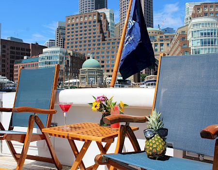 Two lounge chairs, a small table with cocktails and flowers, and a pineapple wearing sunglasses on a boat, with city buildings in the background.