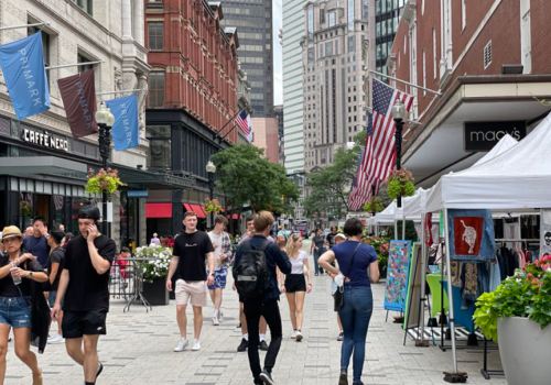 People are walking in a bustling urban street filled with shops, including Macy's, and outdoor vendors, with American flags displayed.