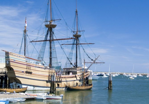 A large wooden sailing ship docked at a harbor with blue skies and several smaller boats in the background.