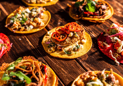 An assortment of colorful tacos with various toppings, including vegetables, meats, and garnishes, arranged on a rustic wooden surface.