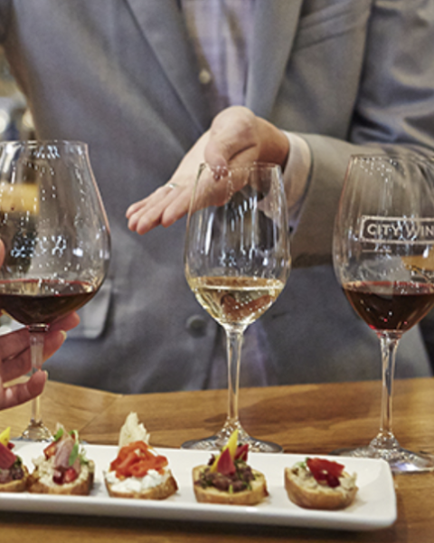 A group of people participating in a wine tasting, holding glasses, with several appetizers on a plate in front of them, ending the sentence.