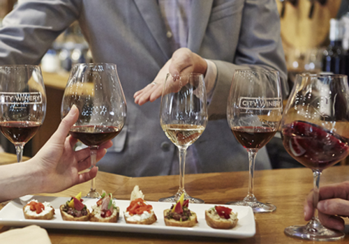 A group of people participating in a wine tasting, holding glasses, with several appetizers on a plate in front of them, ending the sentence.