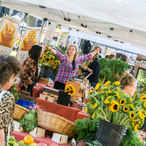 People are shopping at an outdoor market with stalls selling flowers, produce, and other goods under white tents.