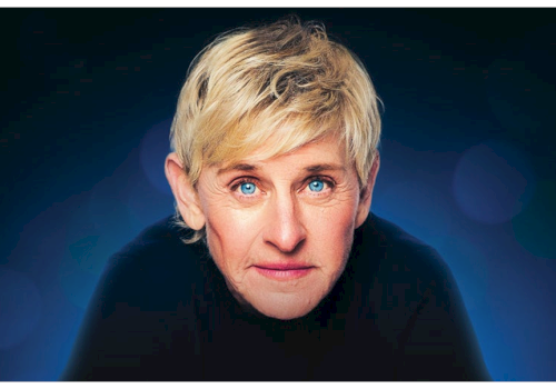 A person with short blonde hair and blue eyes is looking directly at the camera against a blurred dark blue background.