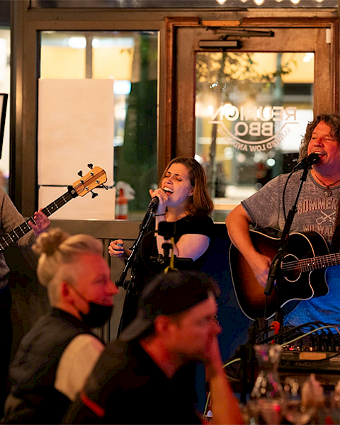 Three musicians perform on a small stage in a lively restaurant or bar, entertaining a crowd of seated patrons.