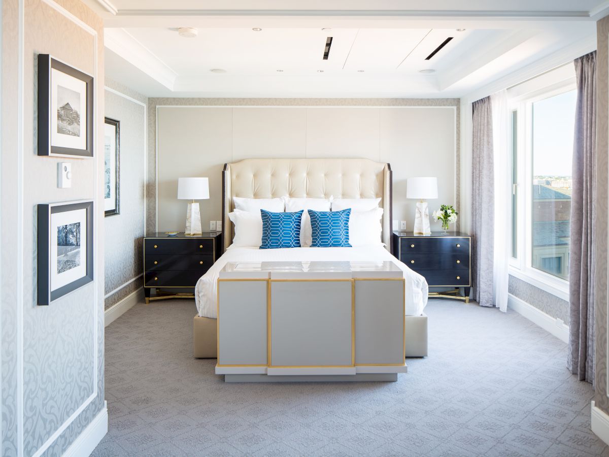 Modern bedroom features a large bed with blue accent pillows, surrounded by nightstands, lamps, framed wall art, and large windows with drapes.