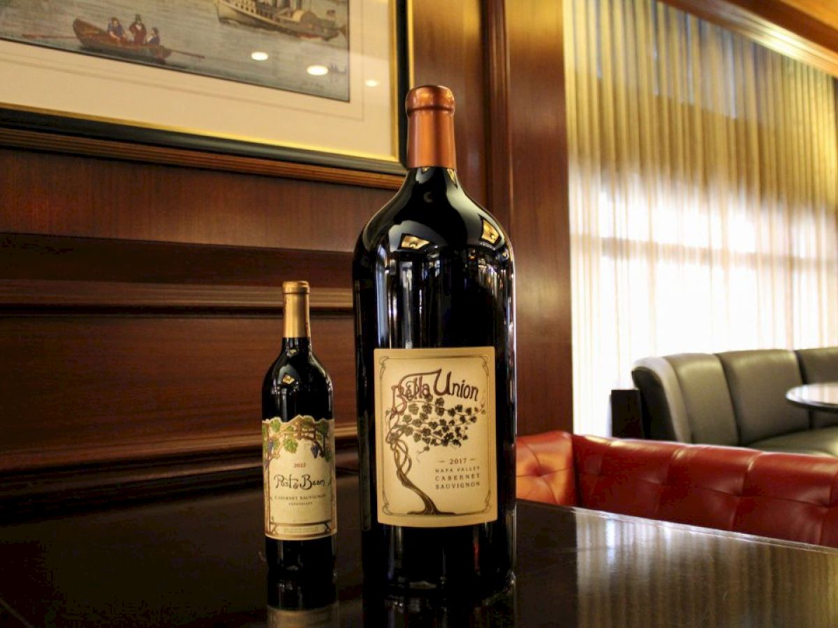 The image shows two bottles of wine, one large and one regular-sized, on a table in a luxurious setting with upholstered seating and a framed artwork.