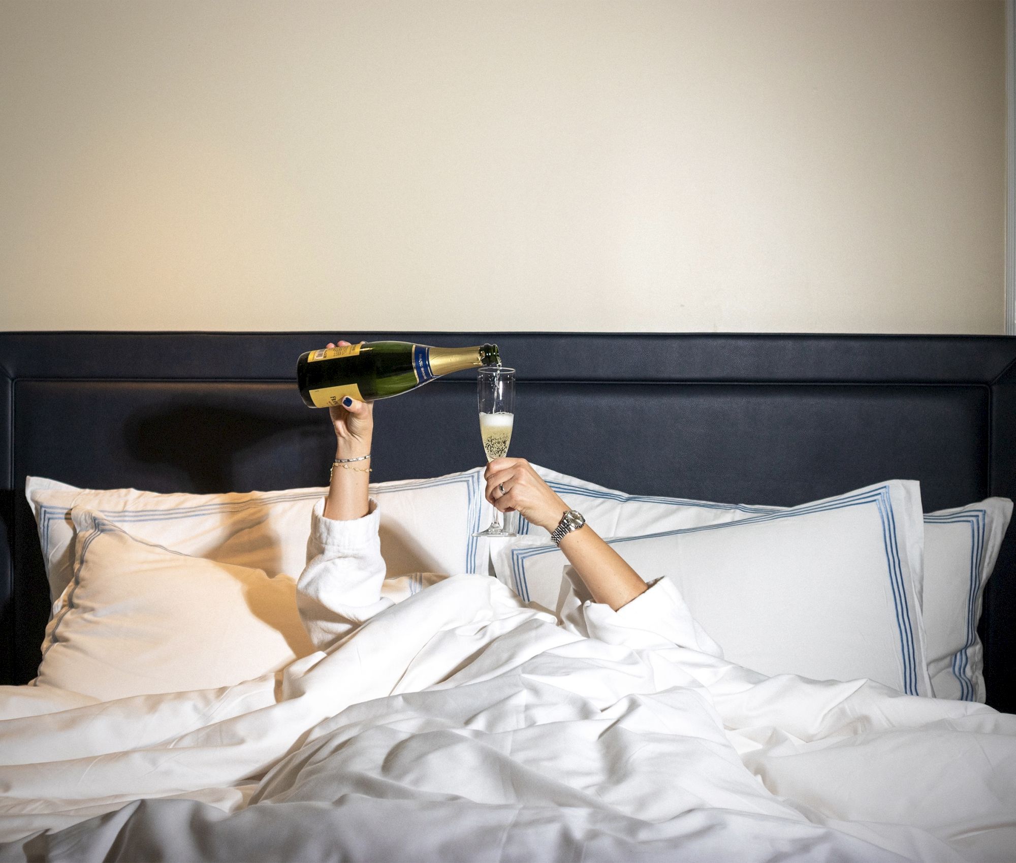 A person lying in bed is pouring champagne into a glass they're holding, surrounded by pillows and white bed linens.