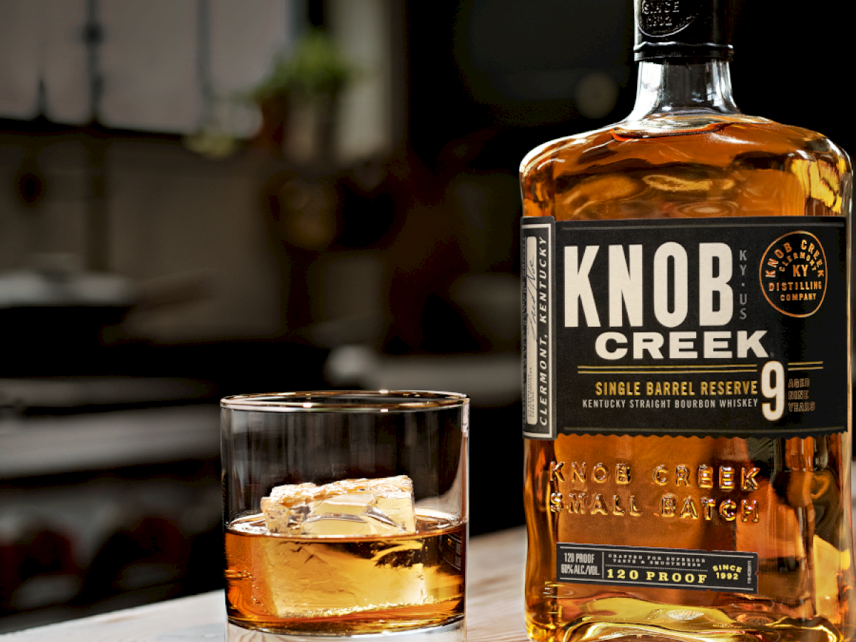 A bottle of Knob Creek whiskey and a glass with whiskey and ice are placed on a wooden table, with a blurred background setting.